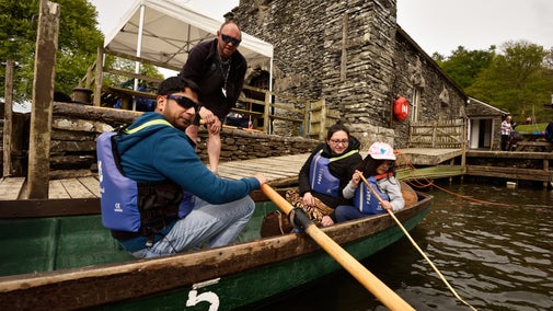 A family sat in a traditional wooden rowboat next to a stone jetty with a stone boathouse in the background.